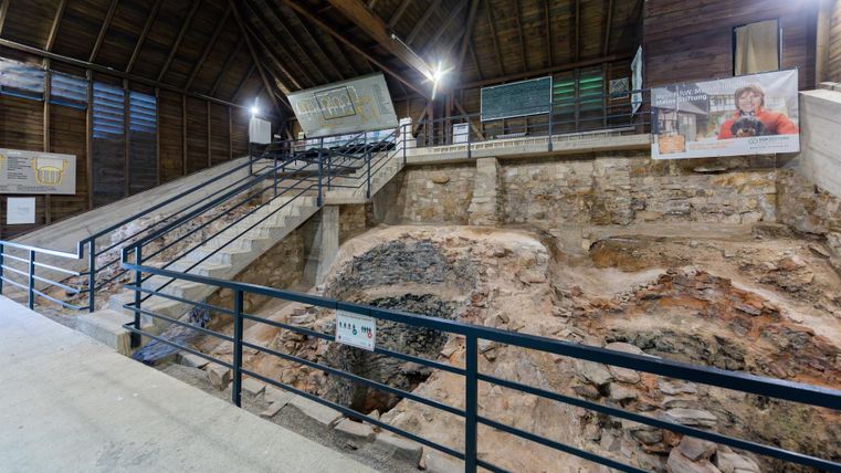 Eine archäologische Ausgrabungsstelle mit Steinrestern und einem darüber liegenden Holzgebäude. Die Umgebung ist besichtigt mit Informationsschildern und einer Treppe.