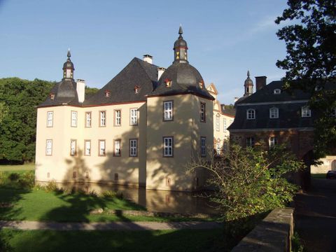 Ein charmantes Schloss mit Türmchen und fensterreicher Fassade, umgeben von Grün. Im Vordergrund ist ein hübscher Garten zu sehen.