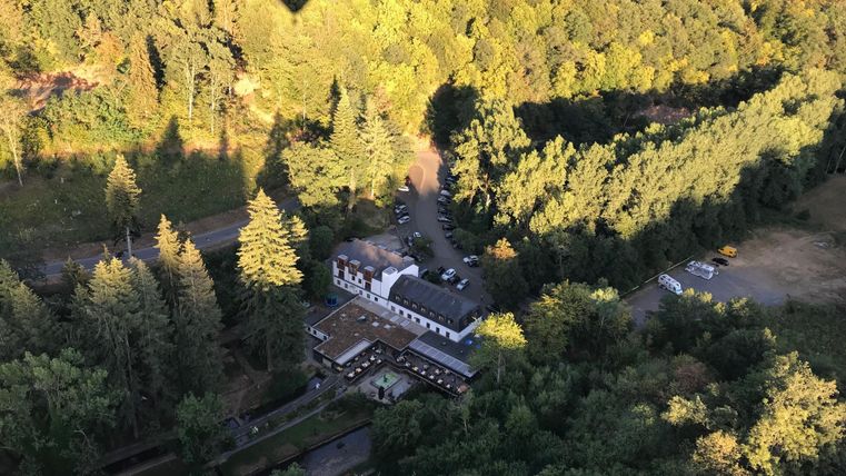 Een luchtfoto van een rustig bosgebied met een gebouw op de voorgrond. Op de achtergrond tekenen zich bomen en de schaduw van een luchtballon af.