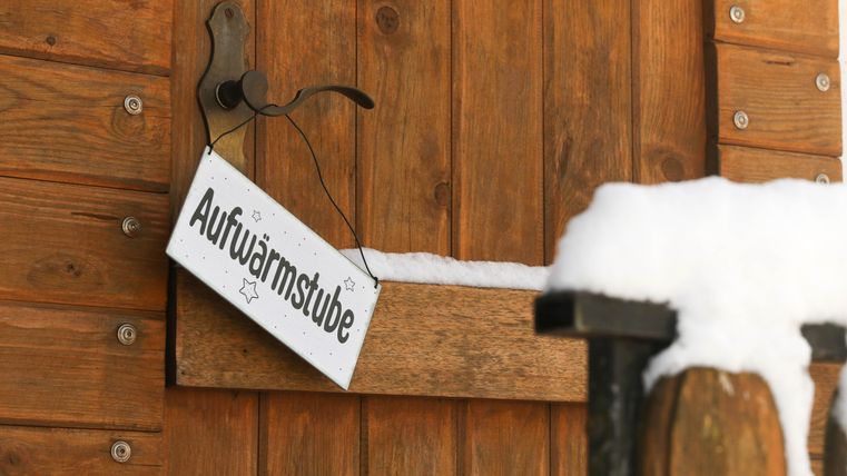 Een houten bord met de inscriptie "Opwarmruimte" hangt aan een deur. Sneeuw ligt op de bovenrand van de deur en op de leuning.