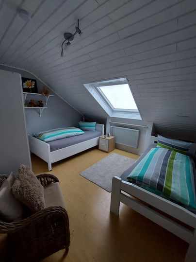 A cozy bedroom with two single beds and a window in the roof. The walls are light, and there is a small table and a chair.