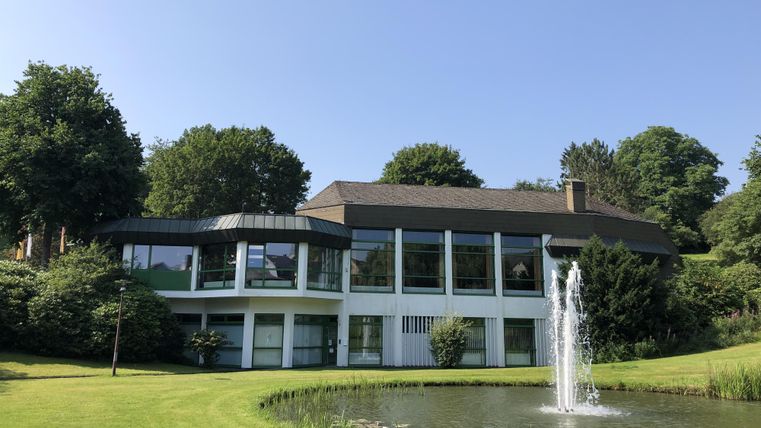 Ein modernes Gebäude mit großen Fenstern und einem schönen Garten. Im Vordergrund befindet sich ein Teich mit einem Sprudel.