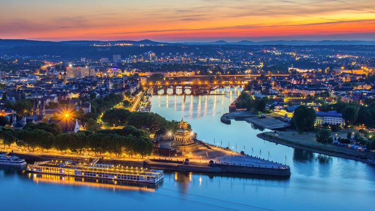 Eine malerische Stadtansicht bei Sonnenuntergang mit einem Fluss, der durch die Lichter der Stadt fließt. Die Farben des Himmels spiegeln sich im Wasser wider.