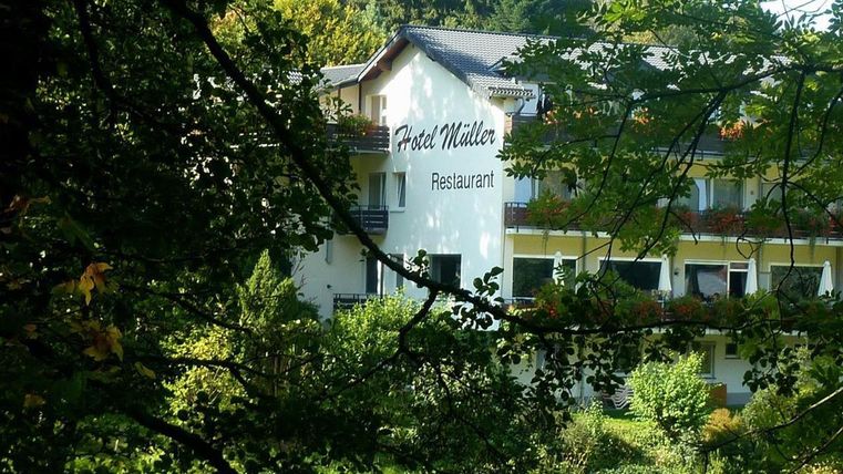 Ein ruhiges Hotel mit dem Namen "Hotel Müller" und einem Restaurant, umgeben von Bäumen und grüner Landschaft. Der Gebäudefront hat Balkone mit Blumen.
