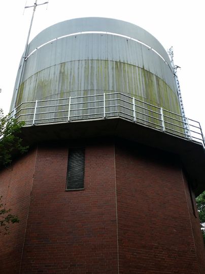A water reservoir with a round, metallic top. The lower part is made of red brick and is surrounded by trees.