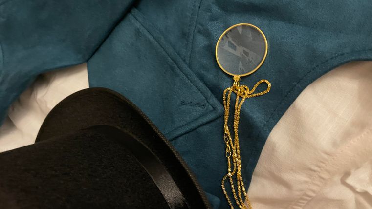 An elegant black top hat lies next to a blue vest. It is accompanied by a golden chain with a round magnifying glass.