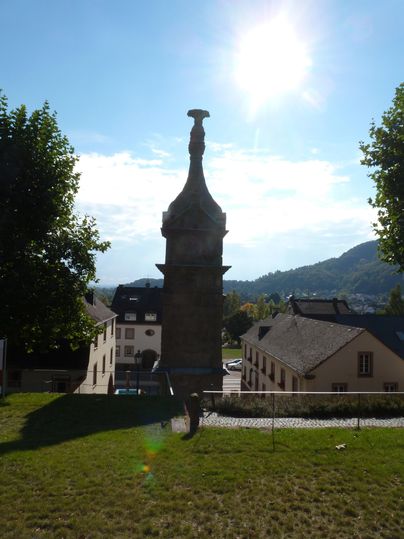 Eine historische Statue mit detaillierten Reliefs steht im Vordergrund. Im Hintergrund sind Bäume und ein klarer blauer Himmel sichtbar.
