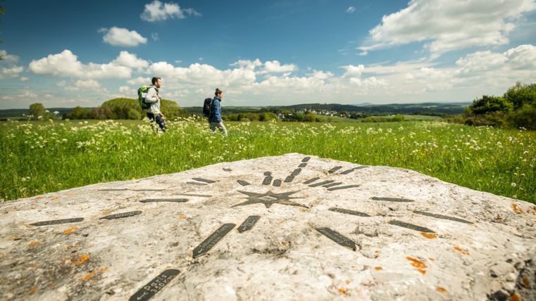 Zwei Wanderer auf einem Pfad durch eine grüne Wiese mit einem großen Stein im Vordergrund, der mit Richtungsmarkierungen versehen ist.