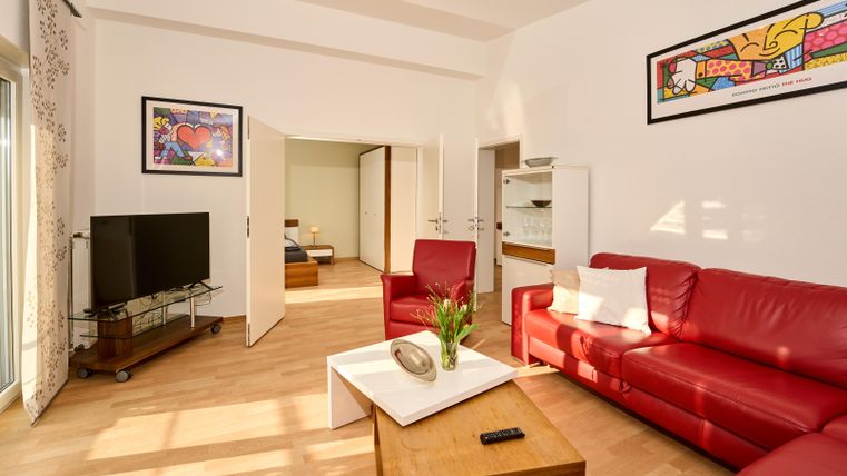 Ein modernes Wohnzimmer mit roten Sofas und einem Kaffeetisch. Im Hintergrund sind ein Fernseher und ein Raum mit einem Bett sichtbar.