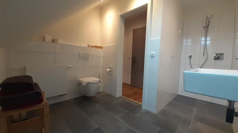 Ein modernes Badezimmer mit grauen Fliesen und hellem Licht. Es gibt eine Dusche, ein Waschbecken und eine Tür, die zu einem anderen Raum führt.