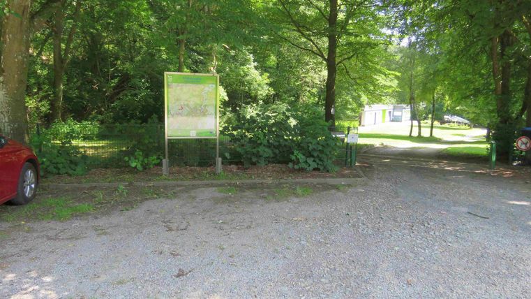 Een smal grindpad tussen bomen leidt naar een parkeerplaats. Op de achtergrond zijn nog meer bomen en een bord zichtbaar.