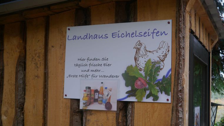 Schild am Landhaus Eichelseifen mit Huhn und Eichelmotiv. Es bietet frische Eier und "Erste Hilfe" für Wanderer an. Ein kleines Bild zeigt diverse Produkte.