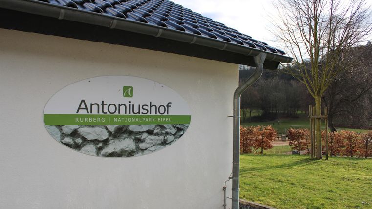 A house with a sign that says "Antoniushof." In the background, trees and fields can be seen.