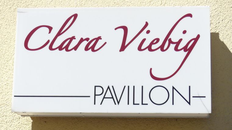 Ein Schild mit dem Namen "Clara Viebig Pavillon". Es ist schlicht gestaltet und hängt an einer Wand.