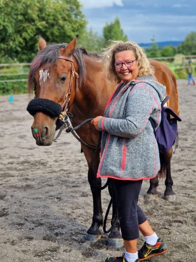 A woman is standing next to a horse in a riding arena. It is a friendly atmosphere with a green background.