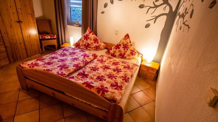 A cozy bedroom with a double bed and colorful floral patterns on the bedding. On the wall is a tree motif and there are warm light sources that create an inviting atmosphere.