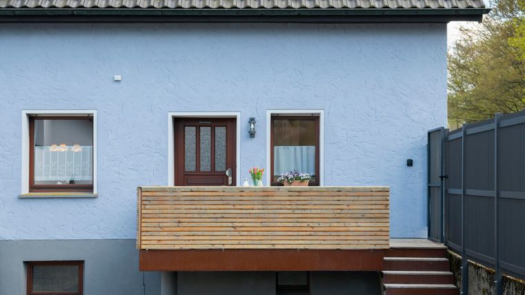 A blue house with a wooden balcony and an entrance door. Flower pots with plants are placed next to the windows.