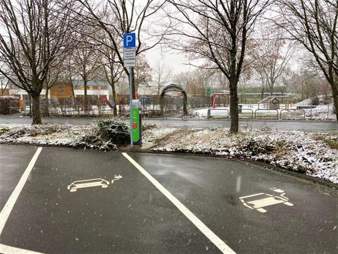 A parking lot with two designated parking spaces and a charging station for electric cars. It is lightly snowing and the ground is covered with a thin layer of snow.