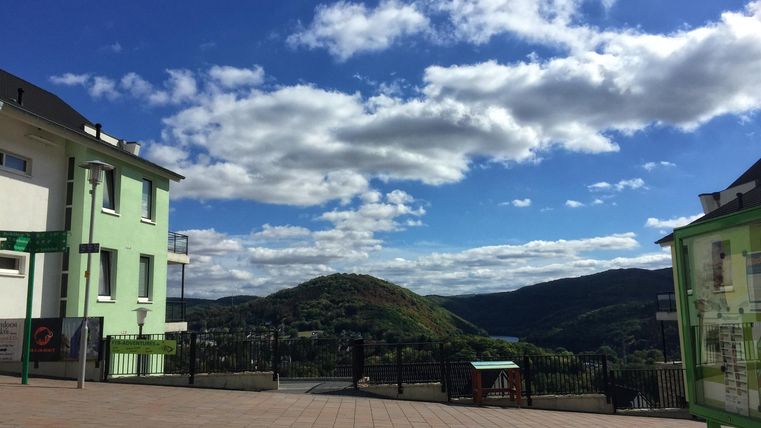 Eine malerische Aussicht auf sanfte Hügel unter einem klaren Himmel mit einigen Wolken. Im Vordergrund sind grüne Gebäude und eine Terrasse sichtbar.