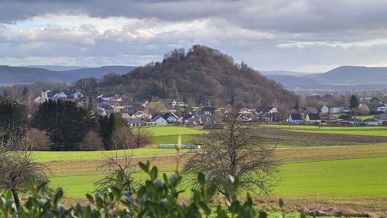 Eine malerische Landschaft mit sanften Hügeln und Wiesen. Im Vordergrund sind Bäume und im Hintergrund sieht man ein kleines Dorf.