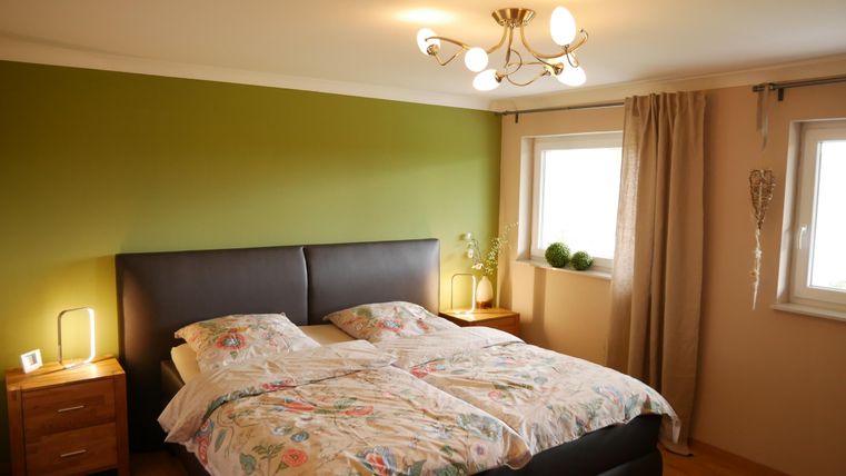 A cozy bedroom with a double bed and colorful bed covers. The walls are in green and beige shades, and the windows let in plenty of daylight.