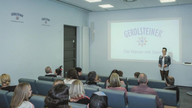 A presentation in the Gerolsteiner Visitor Center. A person is presenting in front of a screen with the Gerolsteiner logo. The audience is sitting in rows.