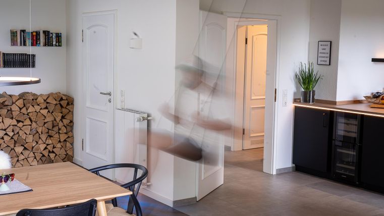 Ein modernes Wohnzimmer mit einem Esstisch und einer Holzwand im Hintergrund. Eine Person bewegt sich schnell, was einen verschwommenen Effekt erzeugt.