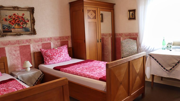 Ein gemütliches Schlafzimmer mit zwei Betten und traditionellen Holzmöbeln. Die Wände sind in sanften Farben gehalten und ein Fenster lässt viel Tageslicht herein.