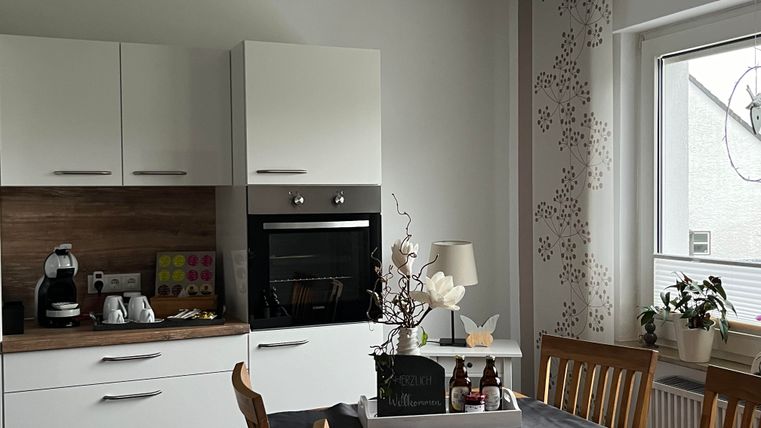 Eine moderne Küche mit einem Holz-Esszimmertisch und einem hellen Raum. Auf der Arbeitsfläche sind Küchengeräte und dekorative Elemente zu sehen.