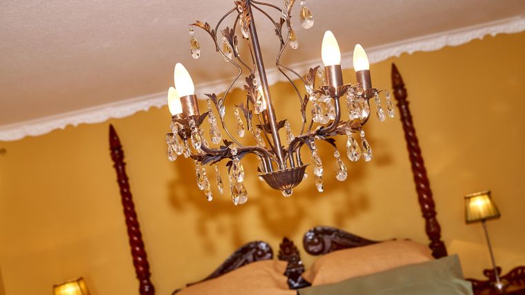 An elegant bedroom with a chandelier and a large bed. The walls are painted in a warm yellow and there is soft lighting from bedside lamps.
