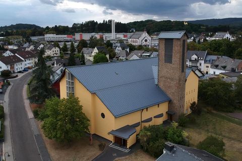 Een moderne kerk met een gele gevel en een hoge toren. Op de achtergrond is een stadslandschap met meerdere gebouwen en heuvels zichtbaar.