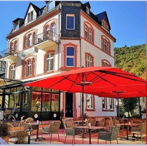 Ein charmantes Café mit einer großen Terrasse und roten Sonnenschirmen. Das Gebäude ist historisch und gut erhalten, umgeben von grüner Natur.