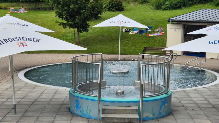 Een klein bubbelbad met water en een ingang van treden. Rondom staan parasols en een groen weiland op de achtergrond.