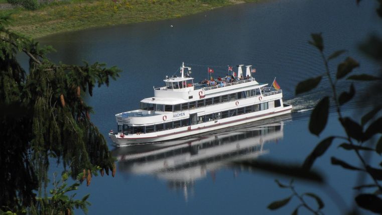 Ein schönes Schiff fährt über einen ruhigen See. Im Hintergrund sind grüne Bäume und eine klare Landschaft zu sehen.