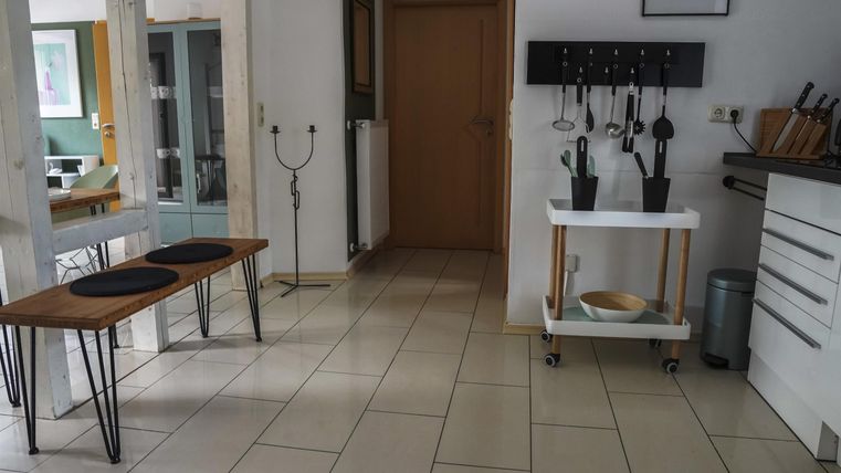 Eine moderne Küche mit hellen Fliesen und funktionaler Einrichtung. Im Vordergrund stehen ein Holztisch und ein Wagen mit Küchenutensilien.