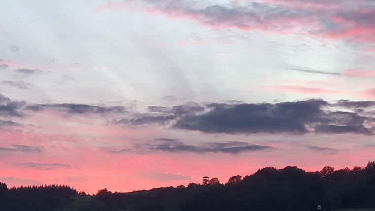 Ein farbenfroher Sonnenuntergang mit rosa und blauen Wolken über einer hügeligen Landschaft. Die Silhouetten von Bäumen sind im Vordergrund sichtbar.