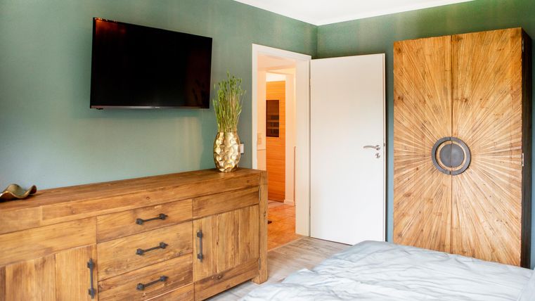 Ein modernes Schlafzimmer mit einer Holzkommode und einem großen Kleiderschrank. An der Wand hängt ein Fernseher, und der Raum hat eine ruhige, grüne Farbgestaltung.