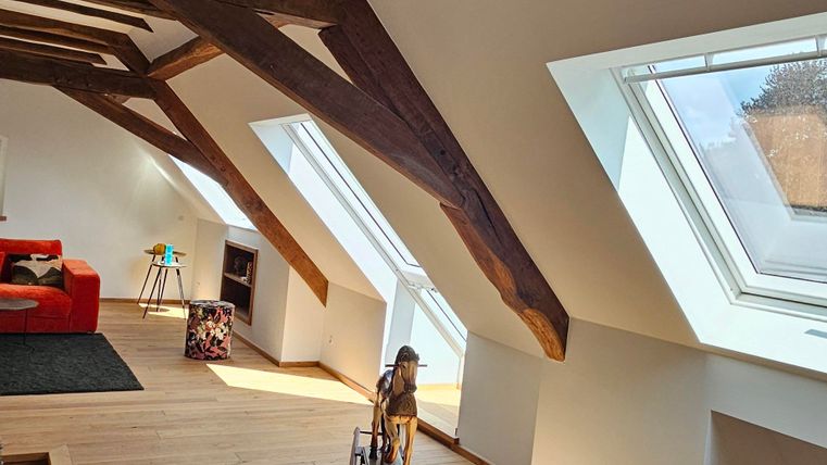A bright room with visible wooden beams and large windows. There is a red sofa and a carpet on the wooden floor.