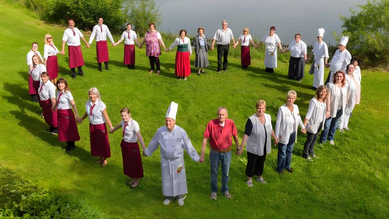 Eine Gruppe von Menschen steht Hand in Hand im Kreis auf einer grünen Wiese. Im Hintergrund ist ein ruhiger See zu sehen.