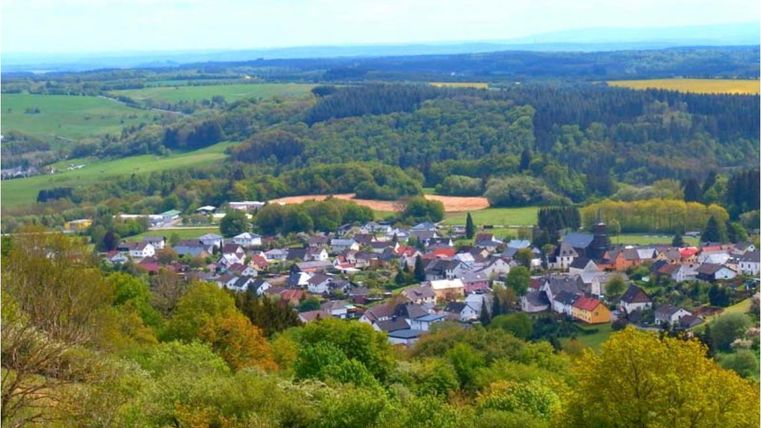 A picturesque view of a small village surrounded by green forests and fields. The sky is clear and the landscape is idyllic.