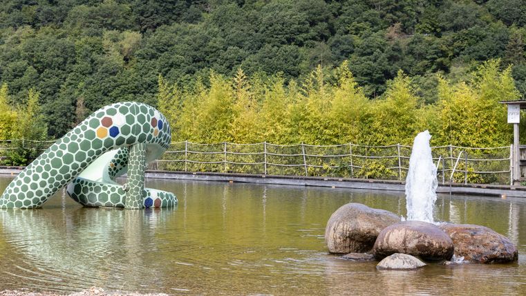Ein bunter Spielplatz mit einer geschwungenen Rutsche und einer Fontäne in einem ruhigen Wasserbecken. Im Hintergrund sind grüne Bäume und Hügel sichtbar.