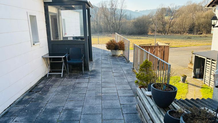 Eine Terrasse mit einem gepflasterten Boden und einem Blick auf die Natur im Hintergrund. Hier stehen einige Pflanzen in Töpfen und ein Stuhl.