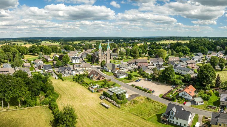 A tranquil landscape with a small village and a church in the center. Surrounding the village are green fields and trees.