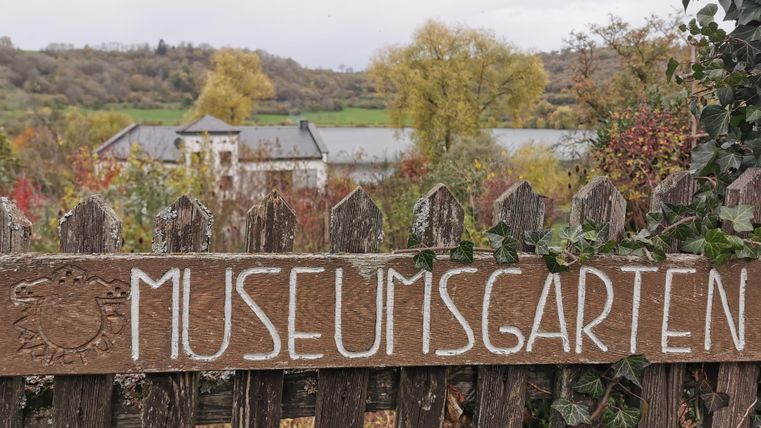 Ein Holzschild mit der Aufschrift "MUSEUMSGARTEN" steht vor einem Garten. Im Hintergrund sind Bäume und ein Gebäude zu sehen.