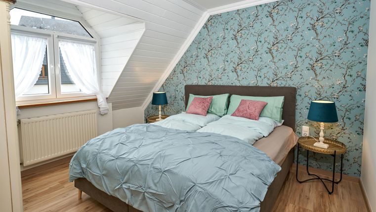 A cozy bedroom with a double bed and colorful cushions. The room has stylish wallpaper and a view from a window.