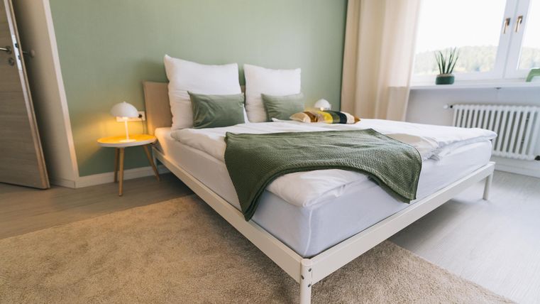 Een moderne slaapkamer met een wit bed en groene kussens. De ruimte heeft een rustige sfeer en veel daglicht.