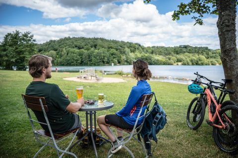 Zwei Personen sitzen an einem Tisch am Wasser und genießen Getränke. Im Hintergrund sind Fahrräder und eine grüne Landschaft zu sehen.
