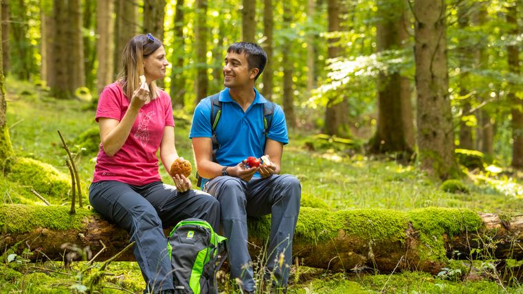 Zwei Personen sitzen auf einem moosbedeckten Baumstamm im Wald und essen.