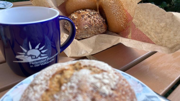 Een kop koffie en verse broodjes op een tafel. Op de achtergrond zijn er meer broodjes zichtbaar in een papieren verpakking.