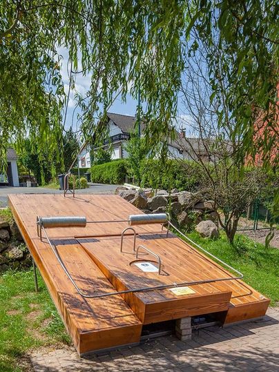 Large wooden construction in the shape of a mouse trap outdoors, surrounded by trees and houses.